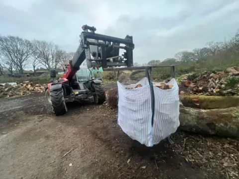 A telehandler lifting a vented bulk bag of split logs in the yard