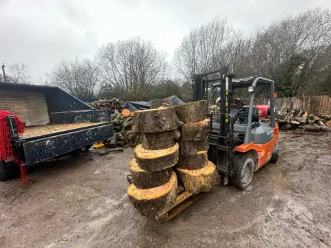 An orange forklift carrying large hardwood rounds across the Woodland Firewood yard